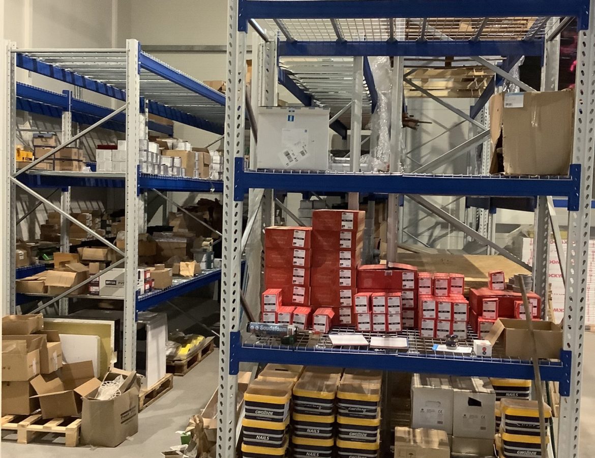 Fragment magazynu z regałami wysokiego składowania wypełnionymi różnorodnymi produktami i paletami na podłodze