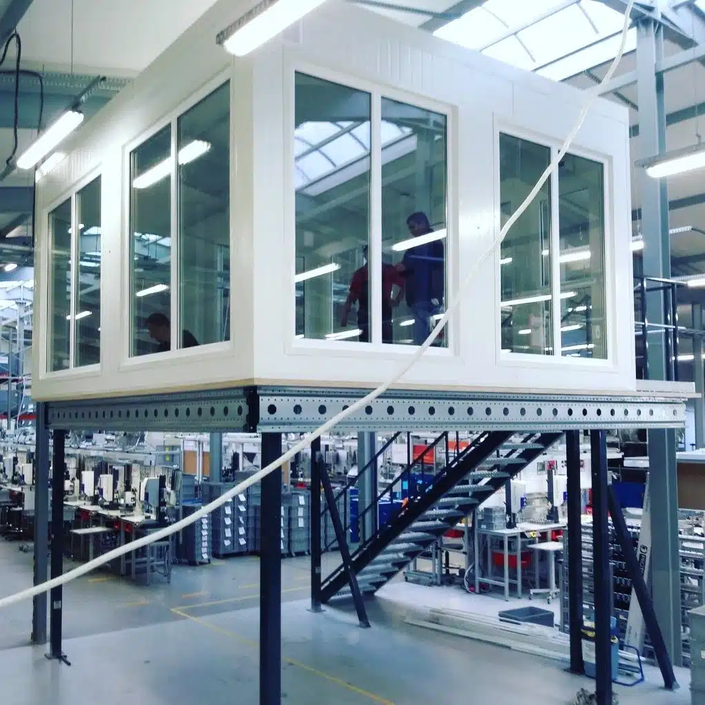 Antresola biurowa w hali produkcyjnej z przeszklonymi ścianami, metalowymi schodami i stanowiskami roboczymi poniżej. Antresola przemysłowa