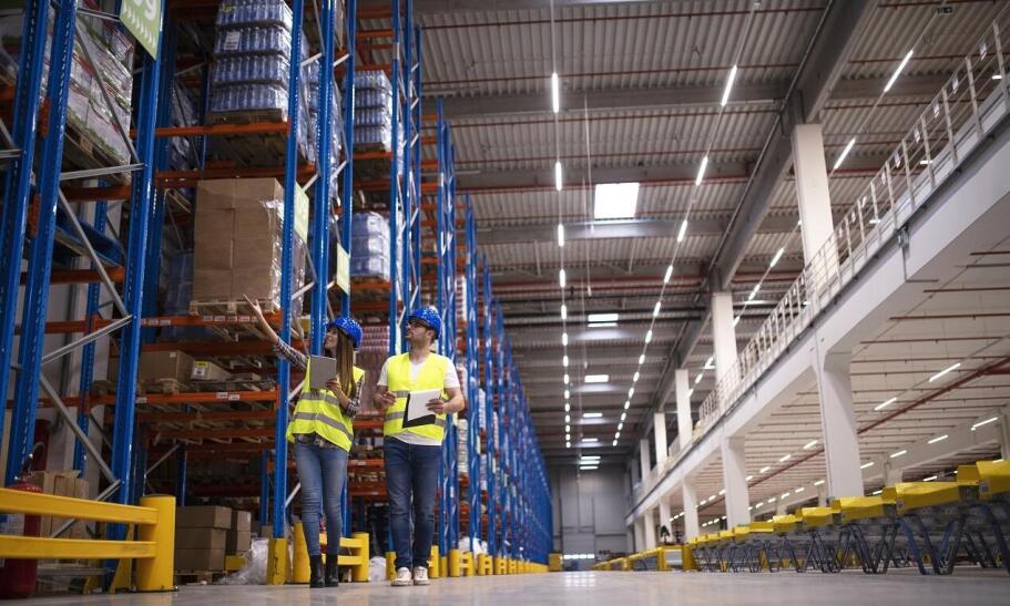 Magazyn logistyczny z pracownikami w żółtych kamizelkach odblaskowych, niebieskich kaskach ochronnych, poruszającymi się między regałami z towarami. Certyfikaty i atesty