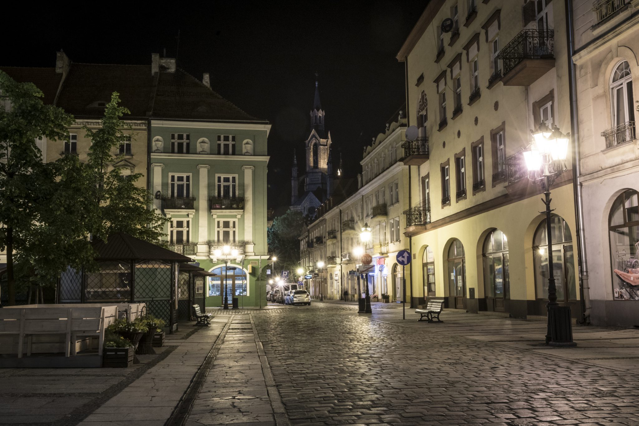 Nocna ulica Śródmiejska w Kaliszu z zabytkową brukarką, kamienicami i wieżą kościoła ewangelickiego w tle, oświetlona stylowymi latarniami. Regały Magazynowe w Kaliszu