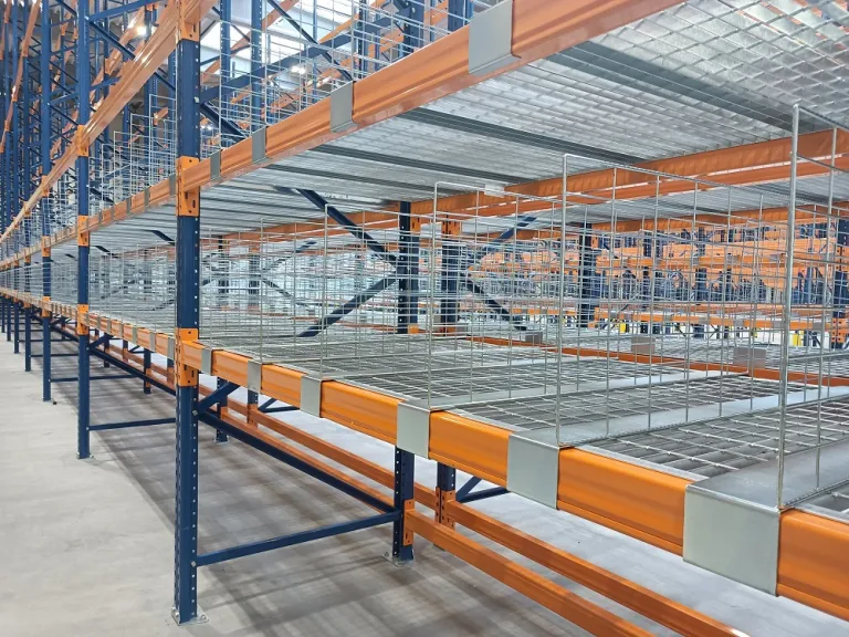 Zdjęcie przedstawia rozległy magazyn z zaawansowanym systemem regałów paletowych. Metalowe konstrukcje w kolorach niebieskim i pomarańczowym sięgają wysoko, tworząc gęstą, uporządkowaną strukturę. Każda półka jest szczelnie wypełniona białymi pudłami i opakowaniami, co świadczy o precyzyjnym zarządzaniu zapasami. Żółte zabezpieczenia u podstawy regałów podkreślają dbałość o bezpieczeństwo. Jasne, mocne oświetlenie LED rozświetla przestrzeń, tworząc wrażenie czystości i profesjonalizmu. Betonowa podłoga oraz stalowe konstrukcje oddają industrialny charakter nowoczesnej logistyki, gdzie liczy się maksymalne wykorzystanie przestrzeni i optymalizacja procesów magazynowych.