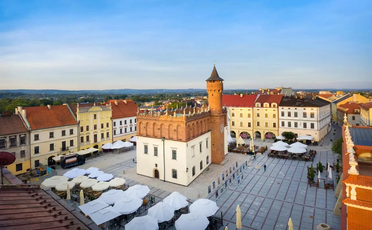 Rynek w Tarnowie z zabytkowym ratuszem, kolorowymi kamienicami i ogródkami restauracyjnymi pod białymi parasolami. Regały Magazynowe w Tarnowie